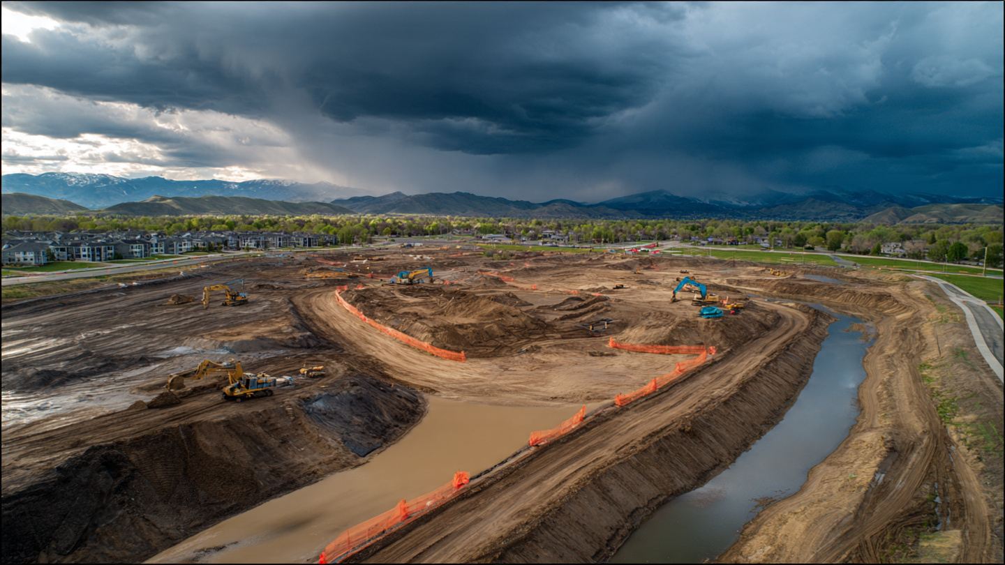 Aerial view of a large active construction site with exposed earth and approaching storm clouds, silt fencing visible along drainage channels